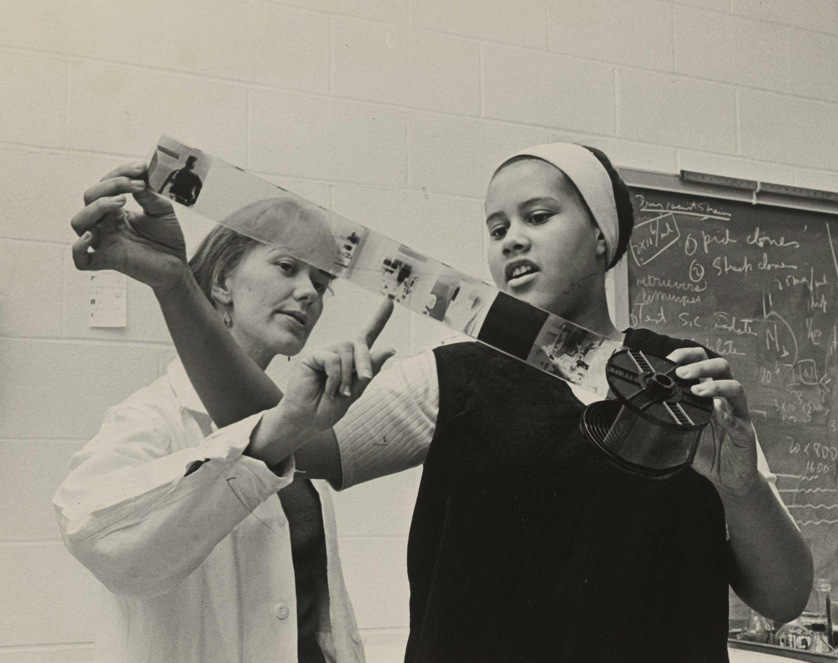 Black and white archival photo of Eva Irene Kataja pointing to a negative on a reel held by Toni Peters.